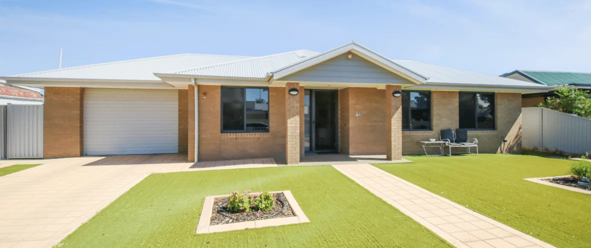 A modern, single-story suburban house in Broken Hill, with a brick exterior and a light grey metal roof. The house features a garage with a white roller door on the left side and a covered accessible entrance with two brick columns. The front yard has artificial grass, a paved walkway leading to the entrance, and a small flower bed with blooming plants. There is a small accessible patio area with outdoor seating on the right side. The sky is clear and blue.