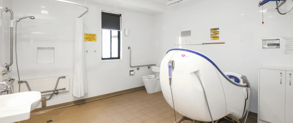 A spacious accessible bathroom with white tiled walls and beige flooring. It features a roll-in shower area with a wall-mounted showerhead, a foldable shower seat, and grab bars. A white sink is mounted on the left side, and a toilet with support rails is positioned near the back. A specialised accessible bathtub with a blue and white design is in the foreground, equipped with handles and an electronic control panel. A ceiling hoist is installed to assist with mobility. A window with a black roller blind provides natural light. Various instructional and safety signs are displayed on the walls.