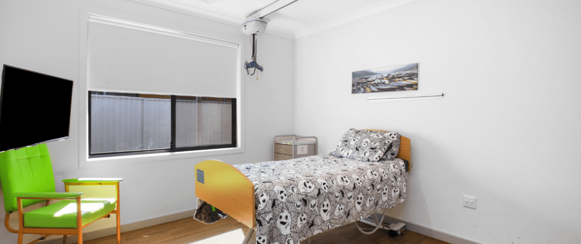 A simple bedroom with a accessible bed featuring a wooden frame and an adjustable base, covered with a black and white cartoon ghost-themed bedspread. The room has white walls, wooden flooring, and a large window with a roller blind. A bright green armchair with wooden legs is positioned next to a small table. A wall-mounted TV is angled towards the bed, and a ceiling hoist is installed. A small set of drawers is next to the bed, and a landscape painting hangs on the wall.