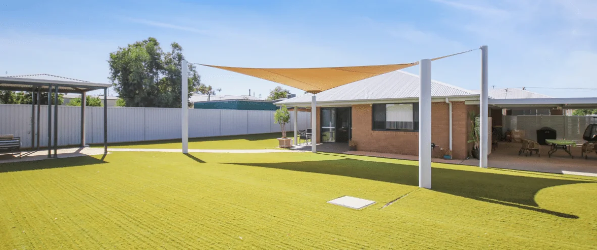 A spacious accessible backyard with artificial grass, featuring a shade sail supported by four white poles. The house has a brick exterior with large windows and a covered patio area with outdoor seating. A gazebo with a metal roof and benches is located to the left, while a barbecue area with a table and chairs is on the right. The yard is enclosed by a tall metal fence, and potted plants add greenery. The sky is clear and blue.