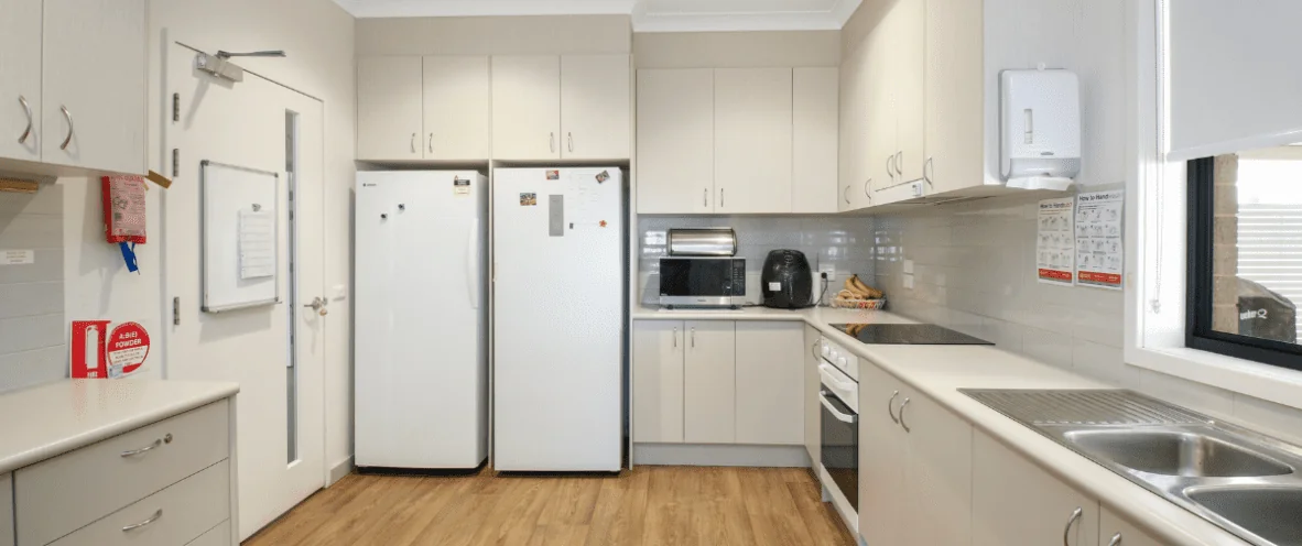 A modern kitchen with beige cabinetry, white countertops, and wooden flooring. The kitchen features two refrigerators, a built-in oven, an electric cooktop, and a stainless steel sink. Small appliances such as a microwave, toaster, and air fryer are placed on the countertop. A fire extinguisher is mounted on the wall near the door, along with safety signs and a whiteboard. A window on the right lets in natural light.