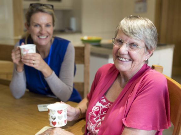 Community care worker joins an an elderly woman for a cup of tea while sitting at her kitchen table.