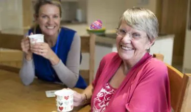 Community care worker joins an an elderly woman for a cup of tea while sitting at her kitchen table.