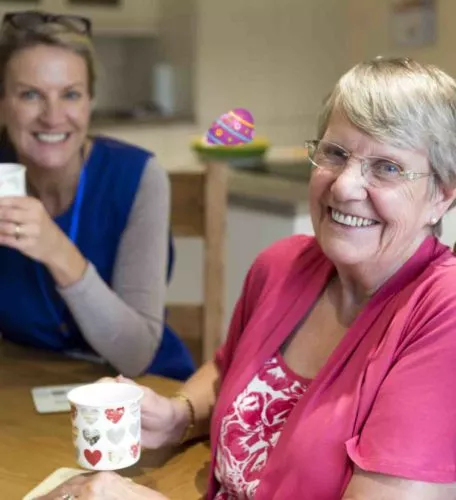 Community care worker joins an an elderly woman for a cup of tea while sitting at her kitchen table.