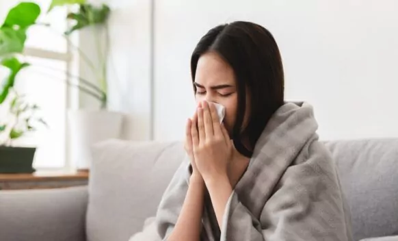 A person with a grey blanket over them. They are blowing their nose with a tissue
