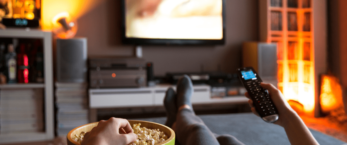 Person sitting on a couch watching television. They have a television remote in one hand and they are eating from a bowl of popcorn with the other hand