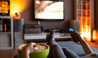 Person sitting on a couch watching television. They have a television remote in one hand and they are eating from a bowl of popcorn with the other hand