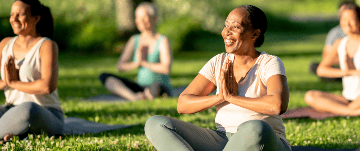 Group of people sitting on yoga mats in a grassy area outside. They have their hands clasped in front of their chests and are smiling