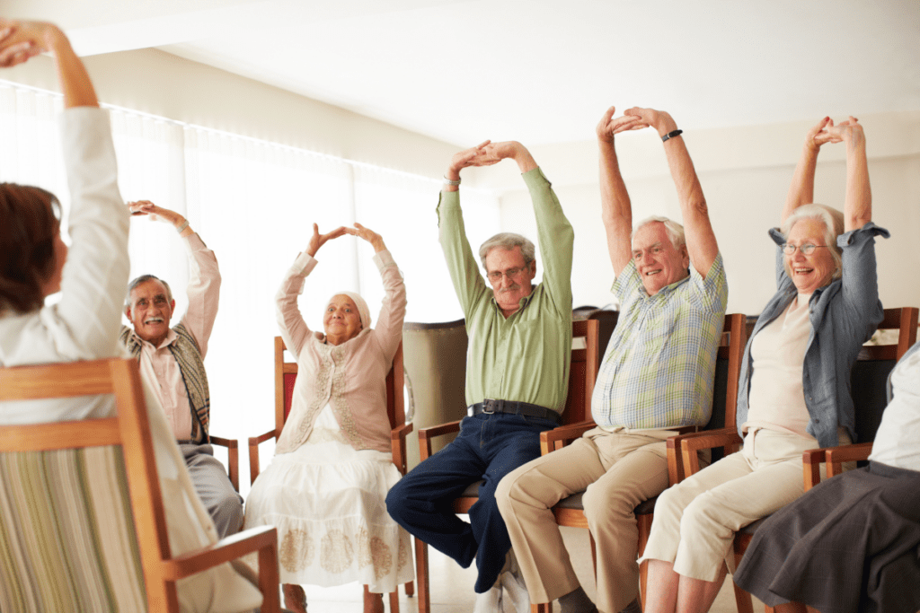 Group of older people performing chair yoga. They all have their hands above their head in a stretch