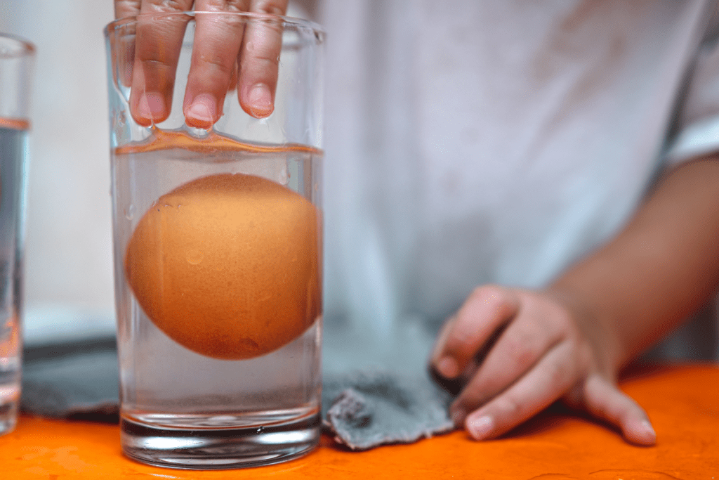 Egg floating in a glass on vinegar. A child is reaching their hand in the jar to pick up the egg
