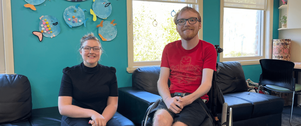 Two people smiling for a photo. One person sitting on a lounge and the other sitting in a wheelchair