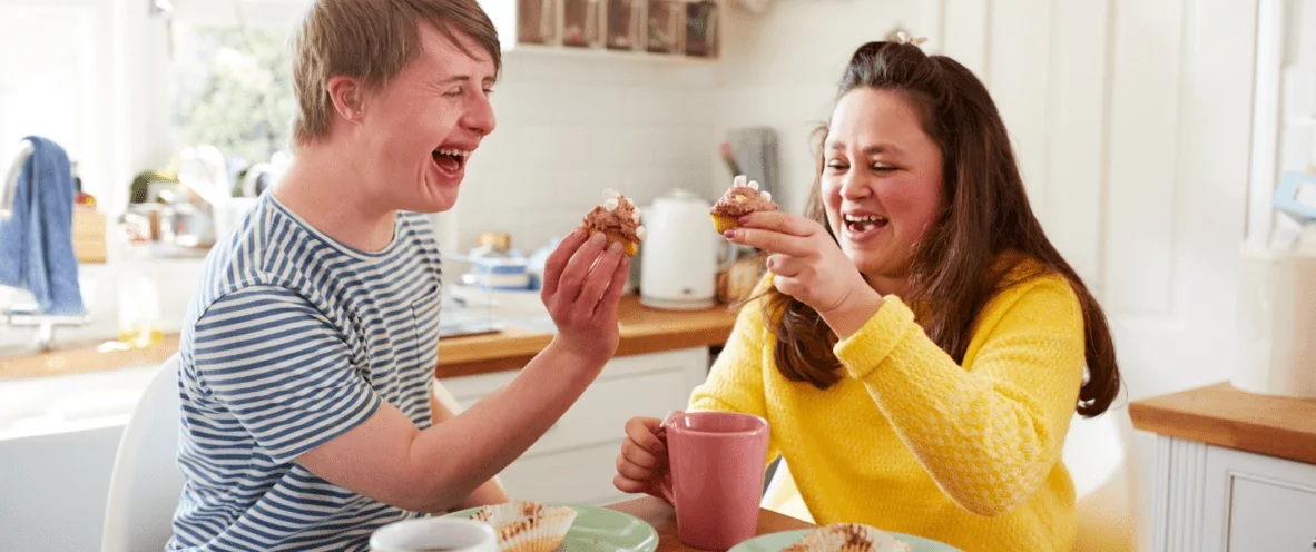 Male and female sitting together at kitchen table laughing and eating cupcakes and drinking tea in SIL