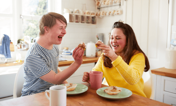 Male and female sitting together at kitchen table laughing and eating cupcakes and drinking tea in SIL