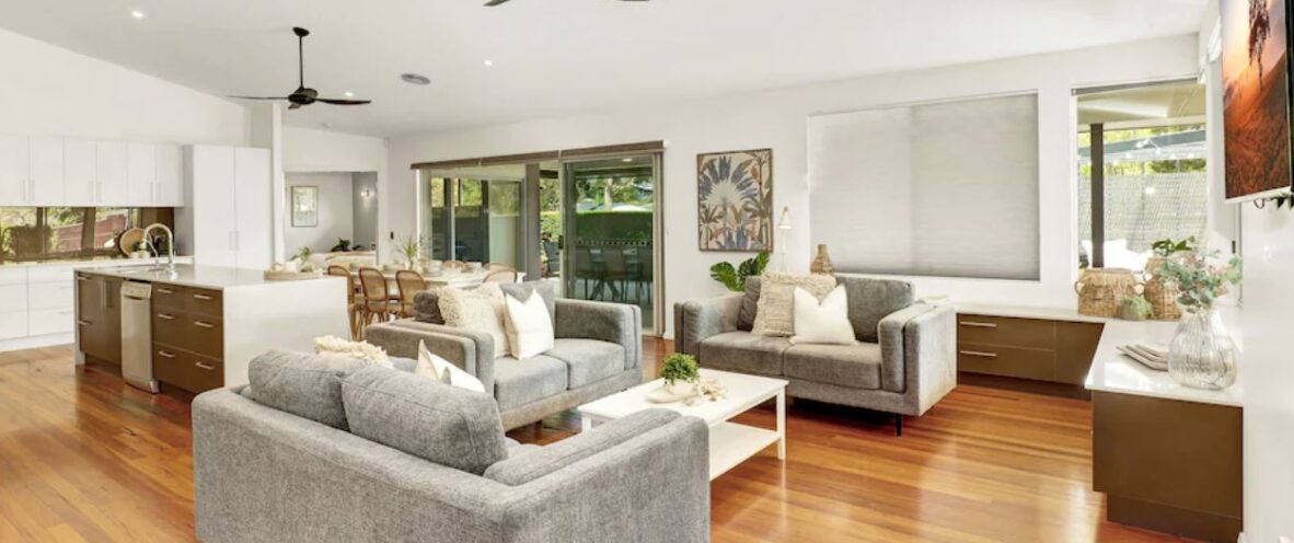 McCabe Coffs Retreat spacious open-plan living area with wooden floors, grey furniture, and modern kitchen. Ceiling fans and large windows with blinds enhance the bright, airy atmosphere.