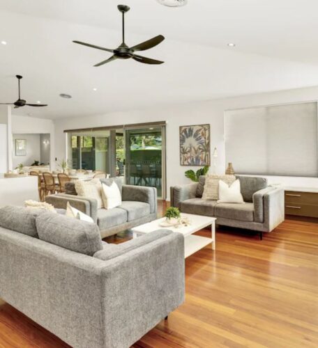 McCabe Coffs Retreat spacious open-plan living area with wooden floors, grey furniture, and modern kitchen. Ceiling fans and large windows with blinds enhance the bright, airy atmosphere.