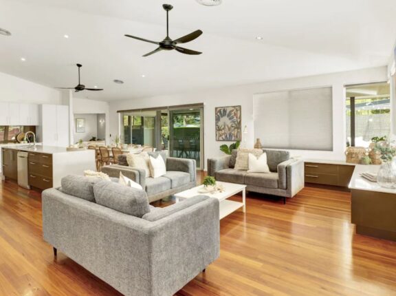 McCabe Coffs Retreat spacious open-plan living area with wooden floors, grey furniture, and modern kitchen. Ceiling fans and large windows with blinds enhance the bright, airy atmosphere.