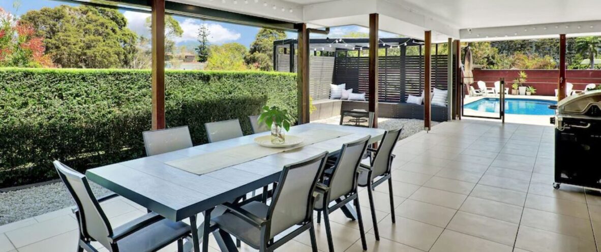 Covered outdoor patio at McCabe Coffs Retreat with dining table and chairs, ceiling fan, view of a green hedge, and pool area in the background.