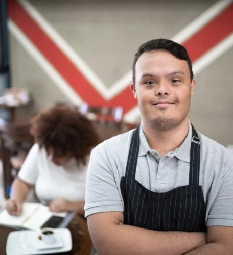 Confident young man with Down syndrome wearing an apron, standing in a café with arms crossed.