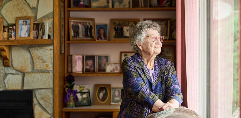 Elderly person with grey hair looks out a window; behind them are shelves and a fireplace filled with framed family photos.