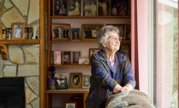 Elderly person with grey hair looks out a window; behind them are shelves and a fireplace filled with framed family photos.