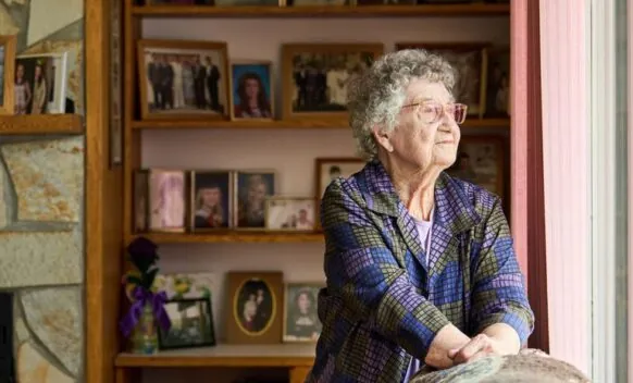 Elderly person with grey hair looks out a window; behind them are shelves and a fireplace filled with framed family photos.